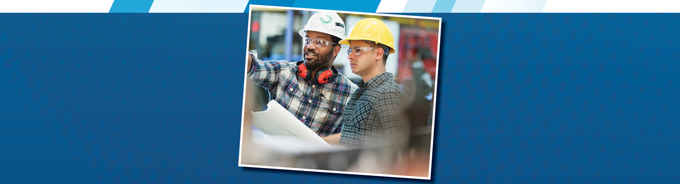 two adults wprking with hardhats and safety goggles. One is holding a blueprint while the other is pointing and explaining something.